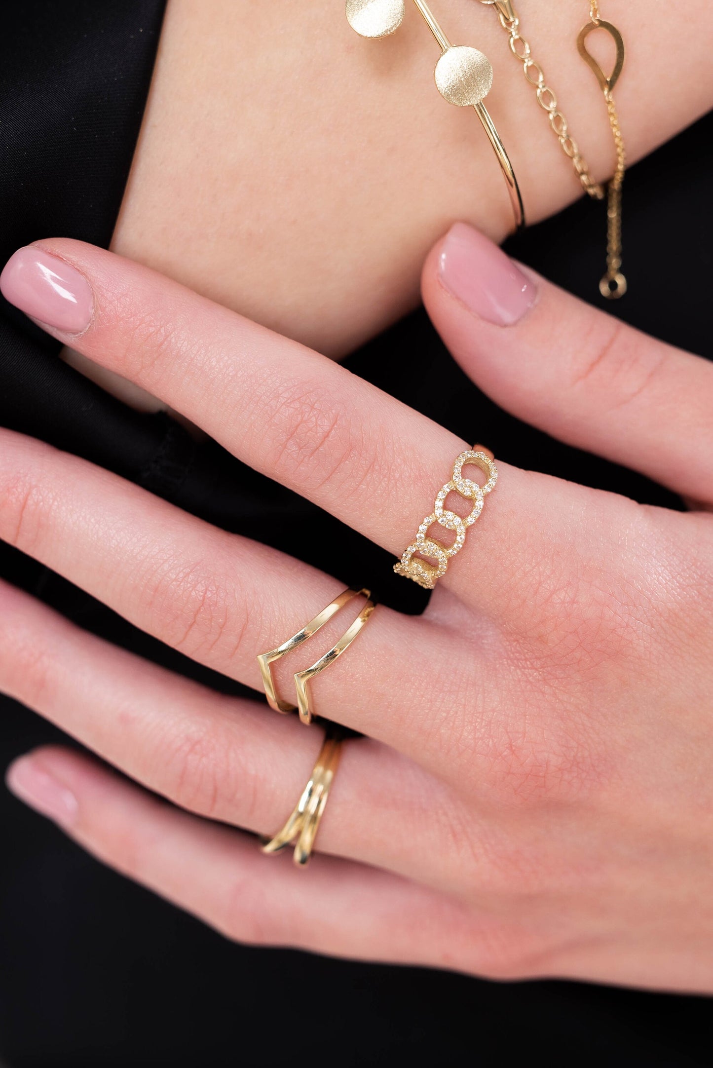 The image showcases a hand adorned with multiple gold rings and bracelets. One ring features a chain-link design with diamonds, while another has a sleek chevron style. The bracelets are minimalist, with one displaying circular disc accents.