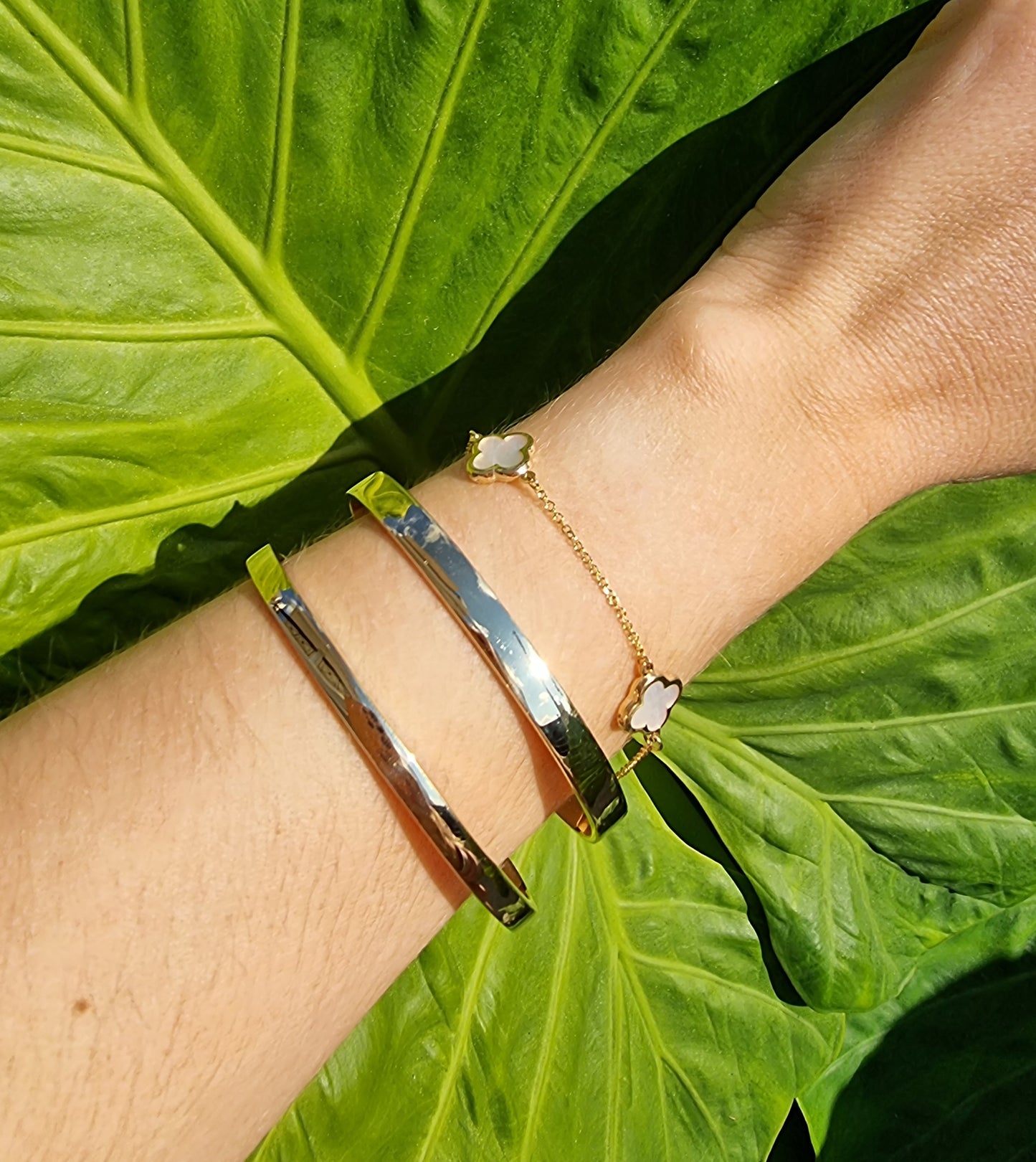 The image depicts a wrist embellished with an assortment of gold bracelets, featuring two solid bangles and a bracelet with a four-leaf clover charm.