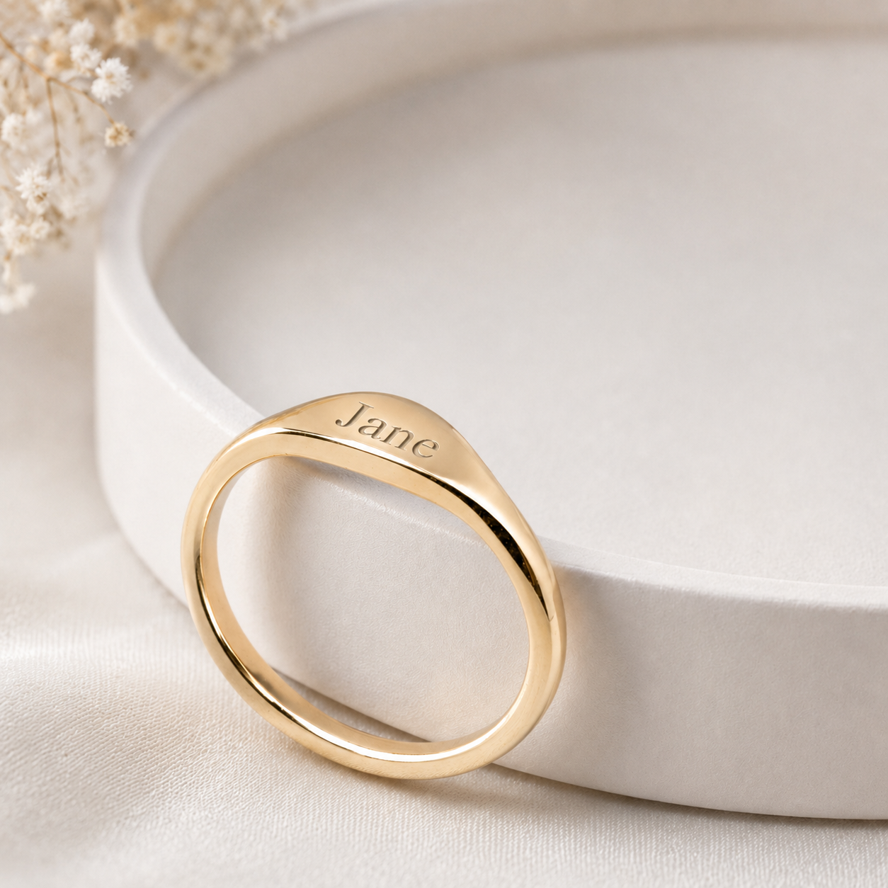 Gold ring with 'jane' engraved on a white surface with dried flowers.