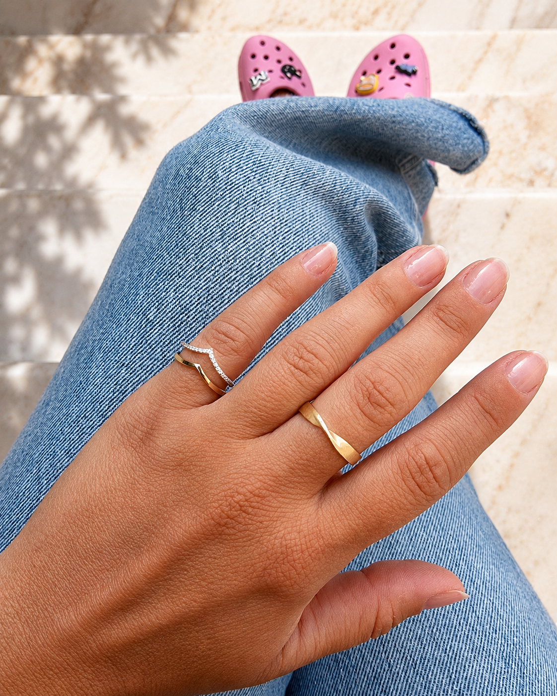 Hand wearing two gold rings with pink Crocs in the background