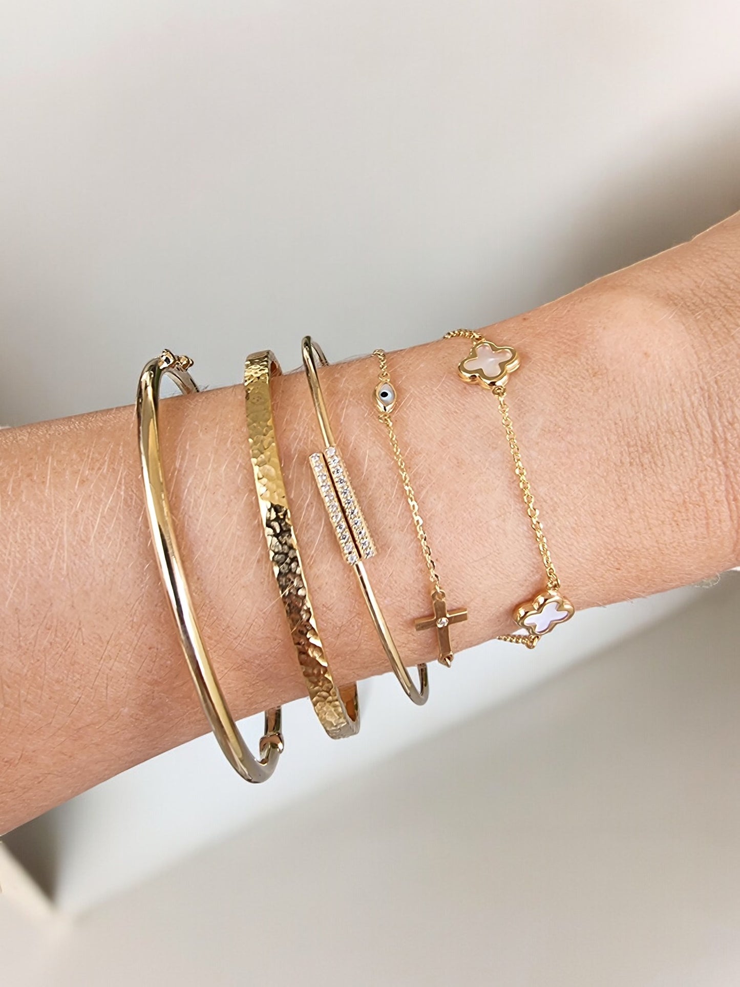 The image shows a wrist adorned with a variety of gold bracelets, including two solid bangles, one with a hammered texture and the other with a smooth finish. There are also delicate chain bracelets featuring small charms