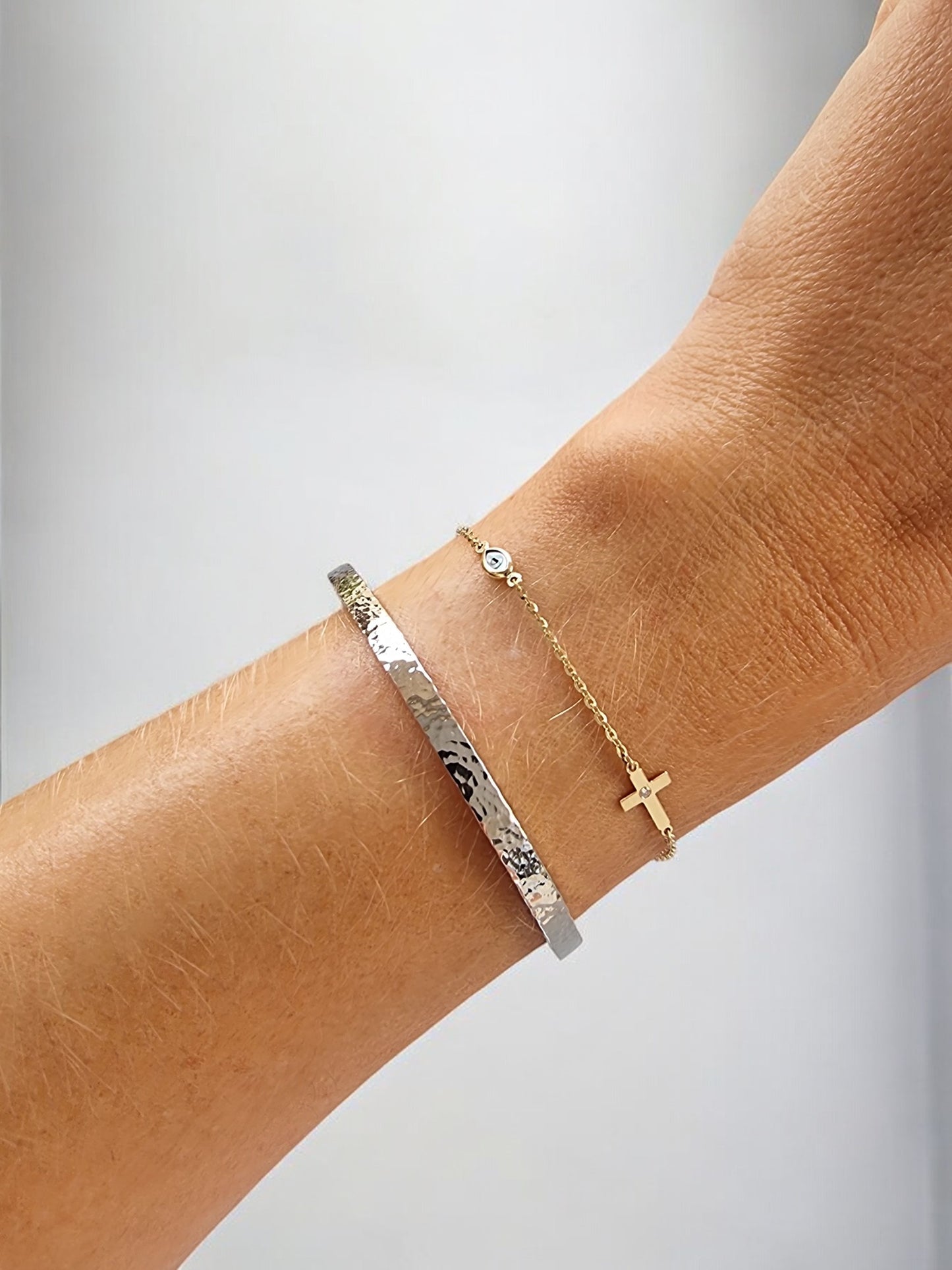 on a hand a 14k yellow gold cross and evil eye bracelet and a hammered white gold bangle bracelet