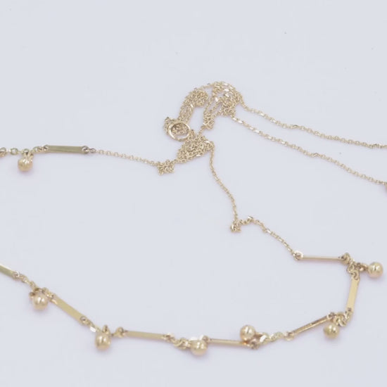  a gold necklace featuring small, shiny, spherical bead-like pendants spaced along a fine chain. Between the beads, there are slim, rectangular bar accents.