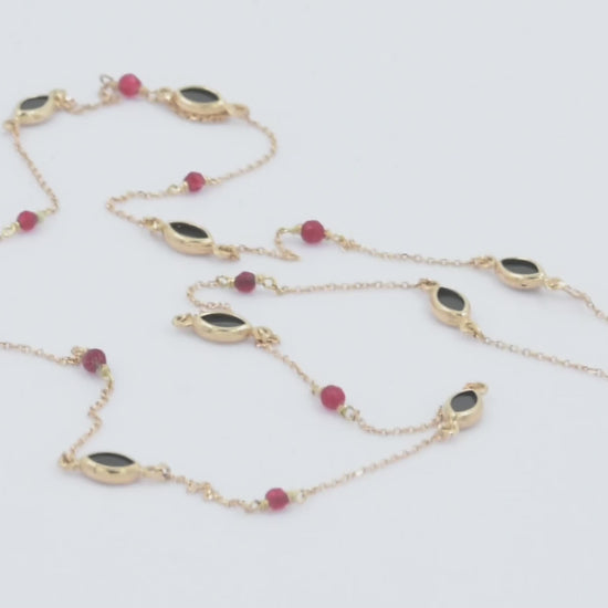 The photo shows a delicate gold necklace featuring small tourmaline gemstones and black marquise-shaped onyx accents, spaced along a fine chain. The contrasting colors create a striking and elegant design