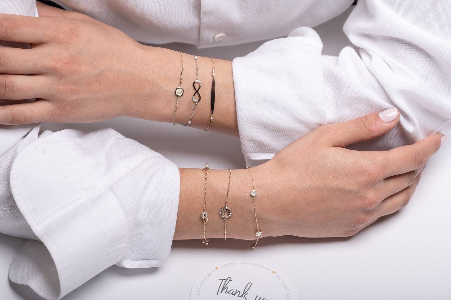 The photo showcases a variety of delicate, layered gold bracelets on a persons wrists. The bracelets feature a mix of elegant designs, including a black infinity symbol, a square bracelet, and sparkling gemstones.