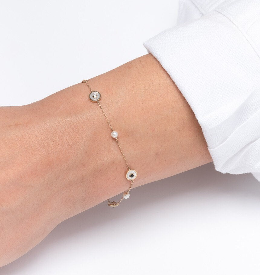 The photo shows a delicate 14k gold station bracelet with circular accents. Each station features alternating black and clear gemstones, set within gold bezels, and interspersed with small pearls along a fine chain.