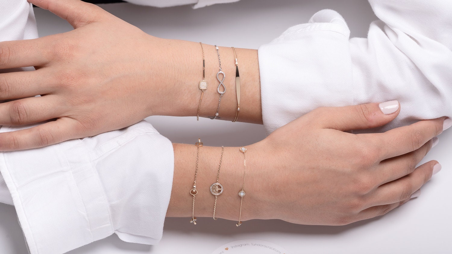 The photo showcases a variety of delicate, layered gold bracelets on a persons wrists. The bracelets feature a mix of elegant designs, including a black infinity symbol, a square bracelet, and sparkling gemstones.