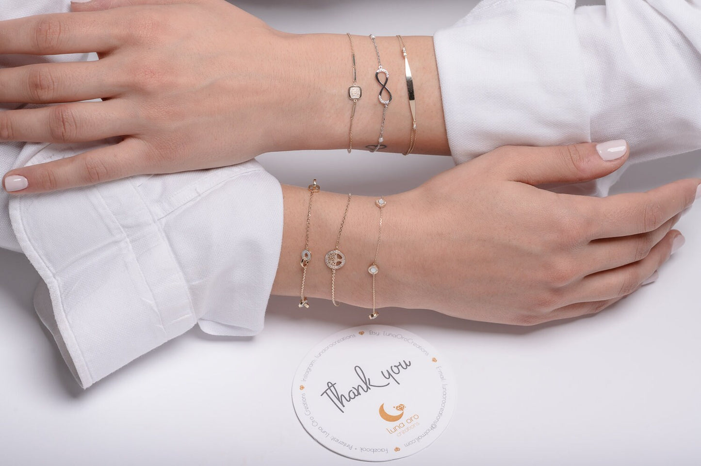 The photo showcases a variety of delicate, layered gold bracelets on a persons wrists. The bracelets feature a mix of elegant designs, including a black infinity symbol, a square bracelet, and sparkling gemstones