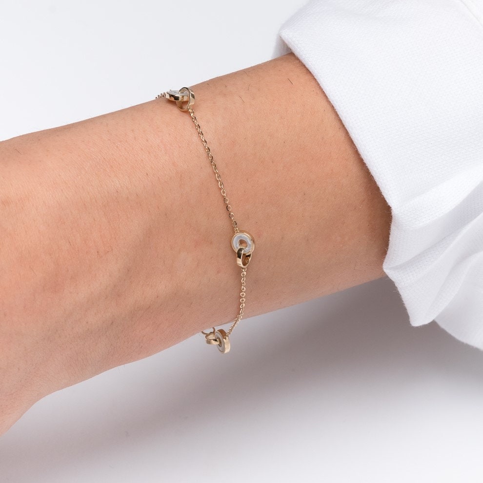 This image shows a delicate gold chain bracelet featuring three interlocking circle motifs. The circles appear to have a blend of gold and mother-of-pearl accents, giving the bracelet an elegant and minimalist look. 