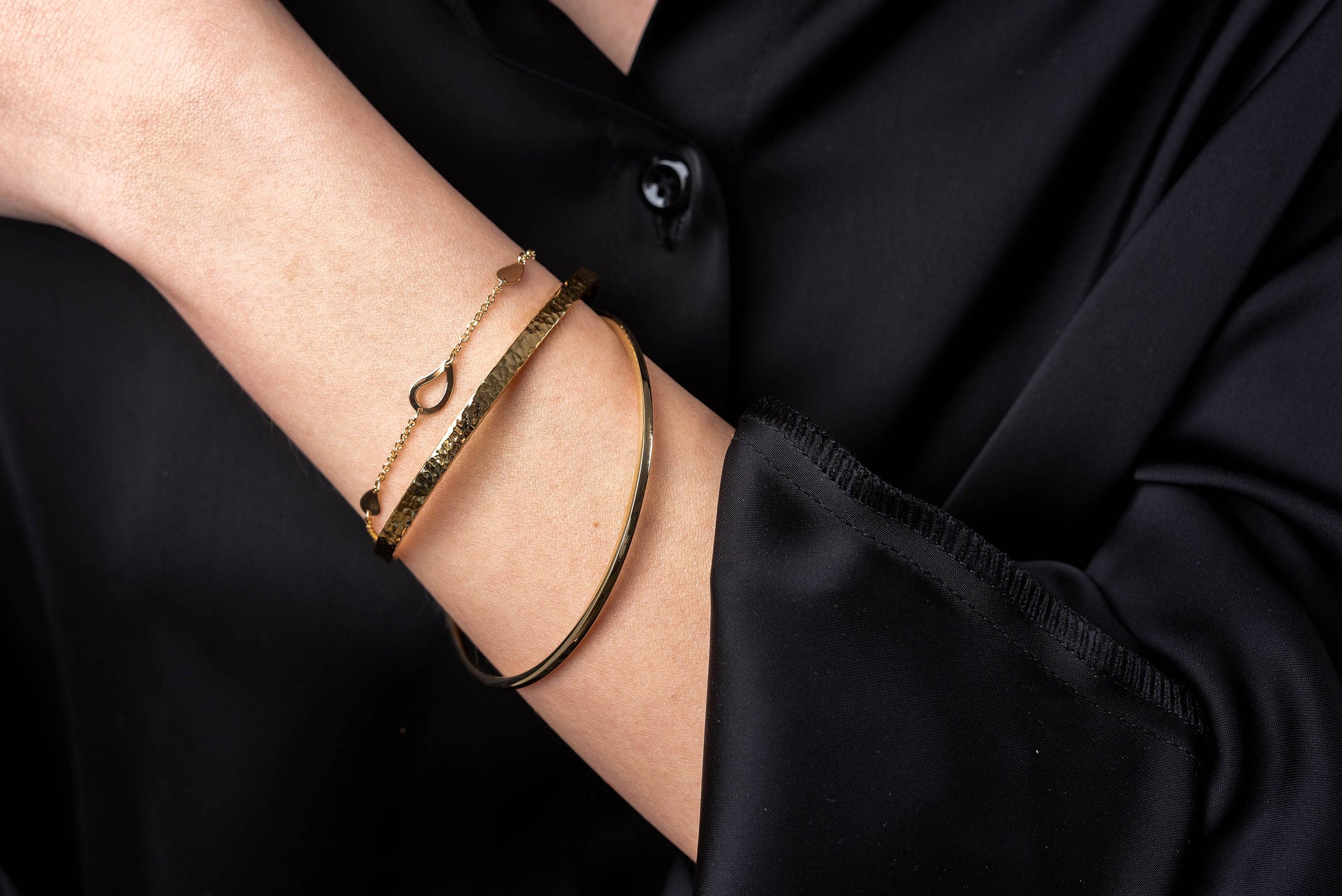 The photo shows a wrist adorned with three gold bracelets: a thin chain bracelet with heart-shaped links, a hammered textured bangle, and a smooth, polished bangle