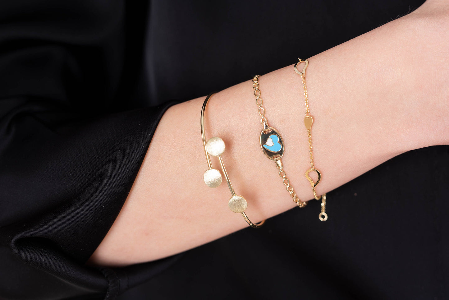 The photo shows a wrist adorned with three delicate gold bracelets. One is an open bangle with three textured round accents, another features a blue and white heart motif in the center, and the third is a fine chain bracelet with leaves 