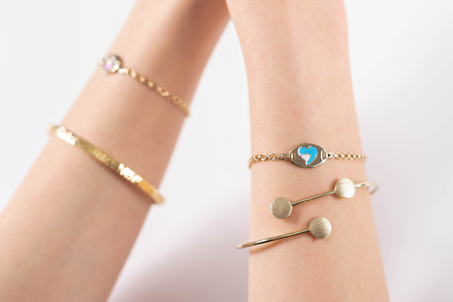 The photo shows a wrist adorned with three delicate gold bracelets. One is an open bangle with three textured round accents, another features a blue and white heart motif in the center, and the third is a fine chain bracelet with leaves 