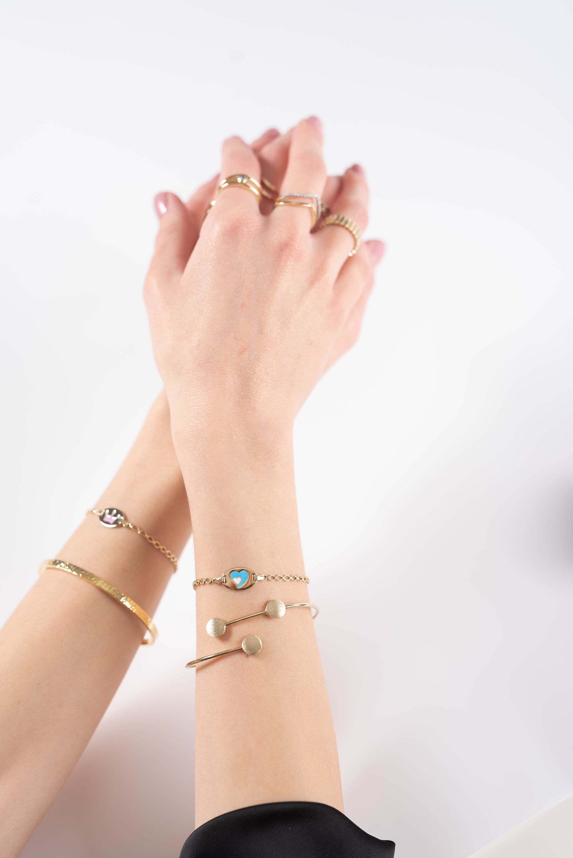 a wrist adorned with three gold bracelets and a hand wearing three gold rings a textured bangle, a chain bracelet with a blue and white heart motif, and a fine chain bracelet with heart-shaped links. The rings are a mix of geometric and chain designs