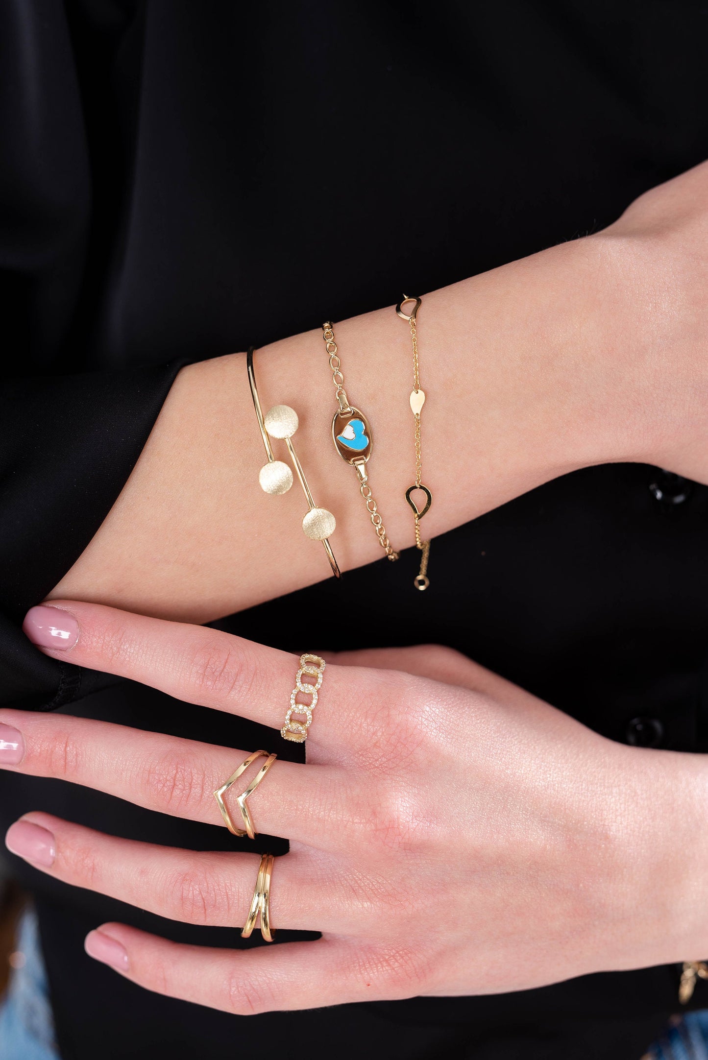 a wrist adorned with three gold bracelets and a hand wearing three gold rings a textured bangle, a chain bracelet with a blue and white heart motif, and a fine chain bracelet with heart-shaped links. The rings are a mix of geometric and chain designs