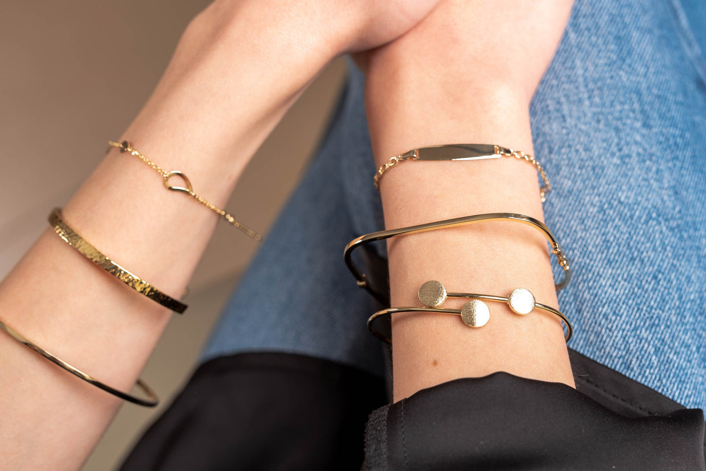 This is an image featuring several gold bracelets on a person's wrist. The bracelets vary in style, including a delicate chain bracelet, a flat bar bracelet, and a couple of minimalist bangle designs.