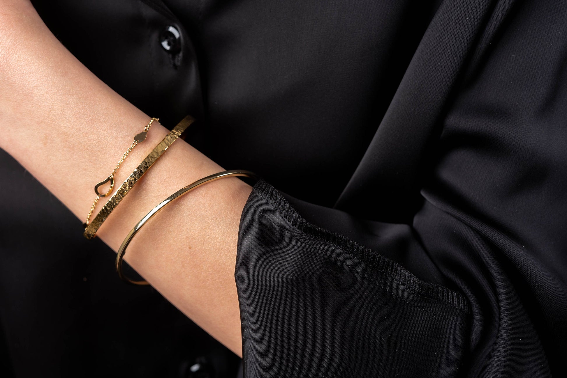 The photo shows a wrist adorned with three gold bracelets: a thin chain bracelet with heart-shaped links, a hammered textured bangle, and a smooth, polished bangle