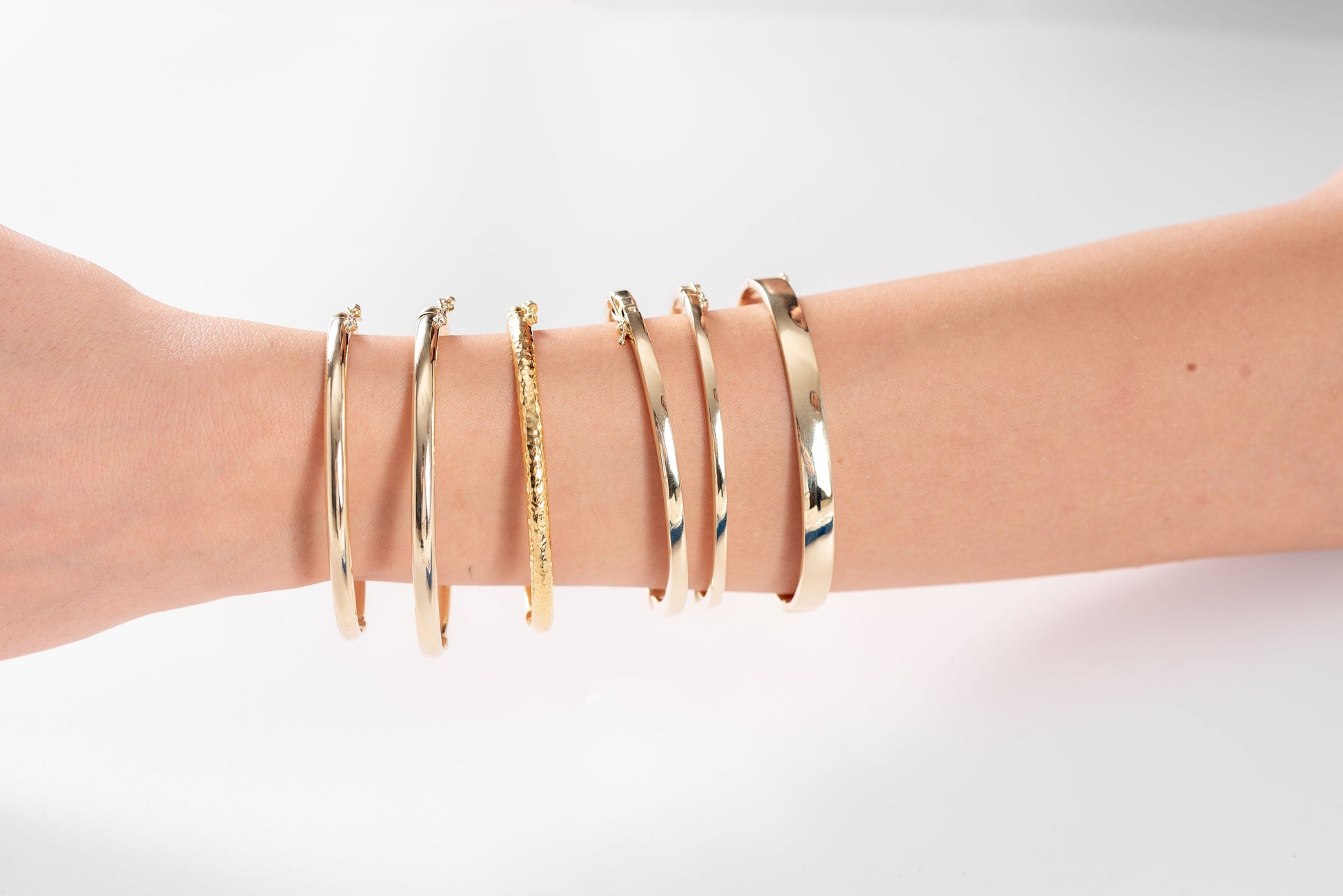 The image shows a wrist adorned with a variety of gold bracelets, including one solid bangle, one with a hammered texture and the other with a smooth finish.