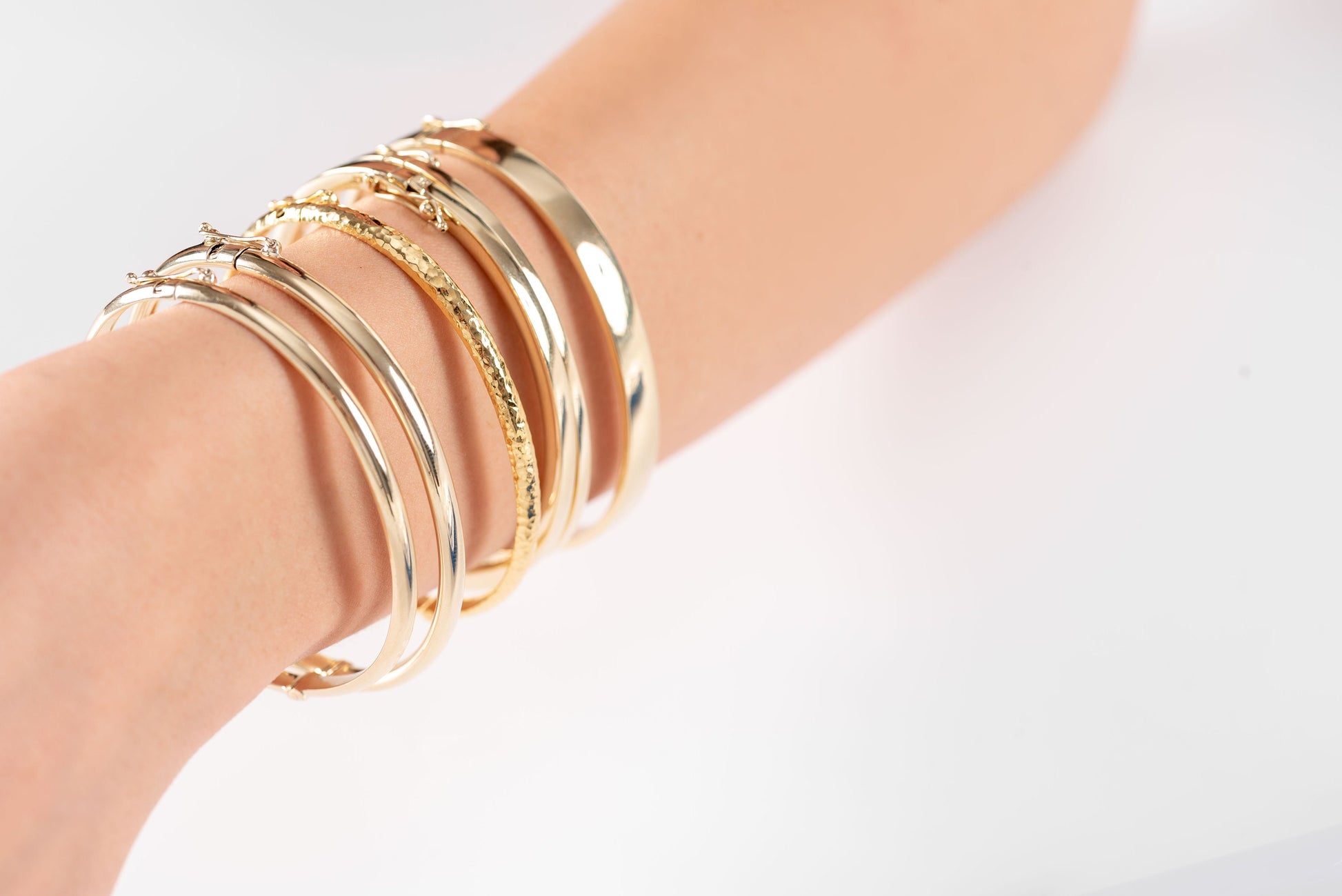 The image shows a wrist adorned with a variety of gold bracelets, including two solid bangles, one with a hammered texture and the other with a smooth finish