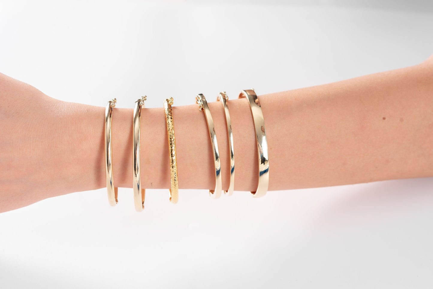 The image shows a wrist adorned with a variety of gold bracelets, including two solid bangles, one with a hammered texture and the other with a smooth finish.