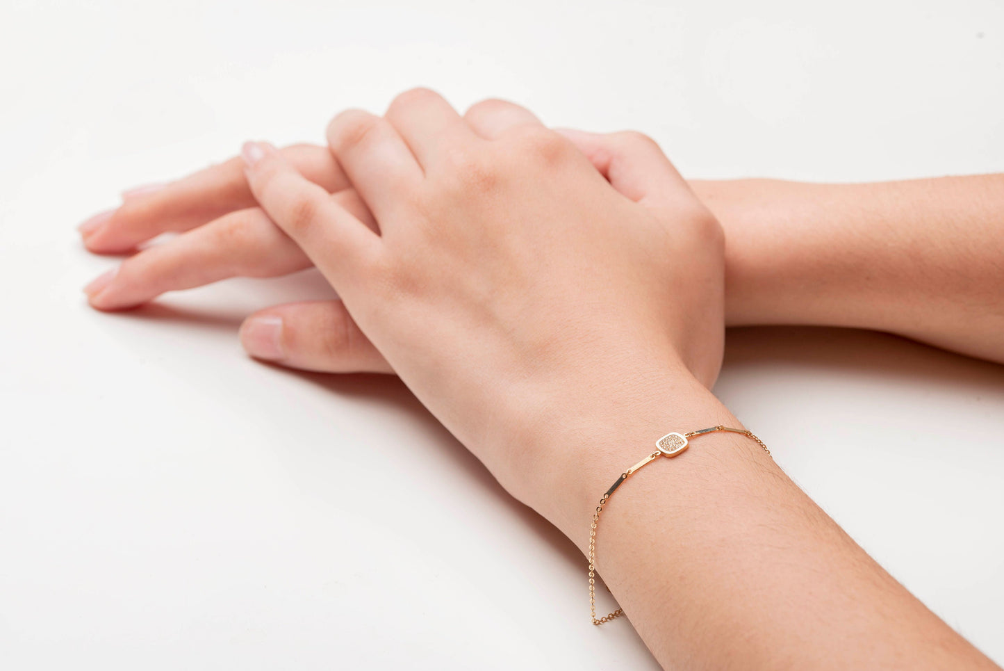 a hand wearing 14k gold charm bracelet with a square charm paved with white cubic zirconia stones