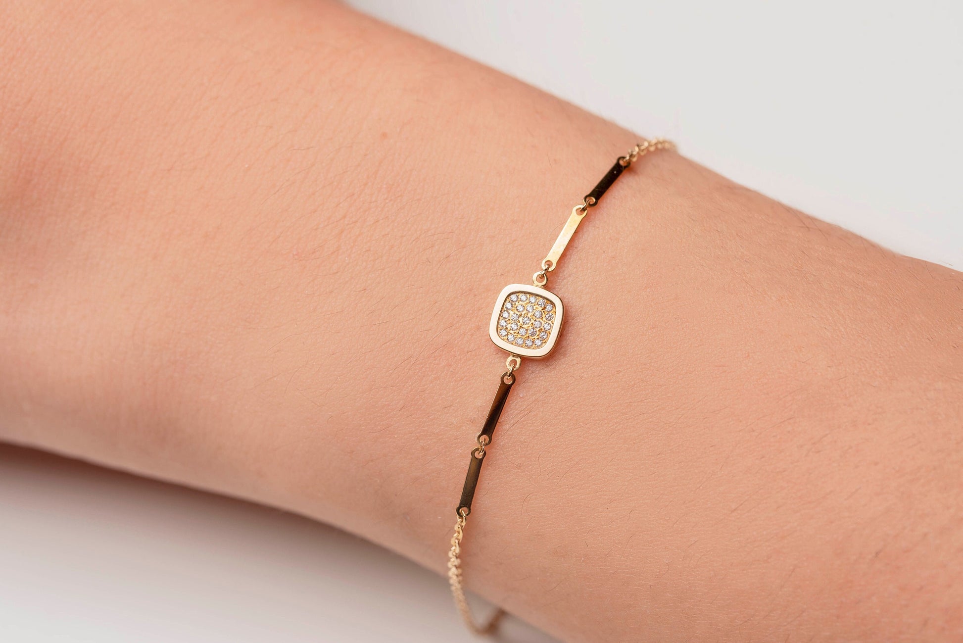 a hand wearing 14k gold charm bracelet with a square charm paved with white cubic zirconia stones