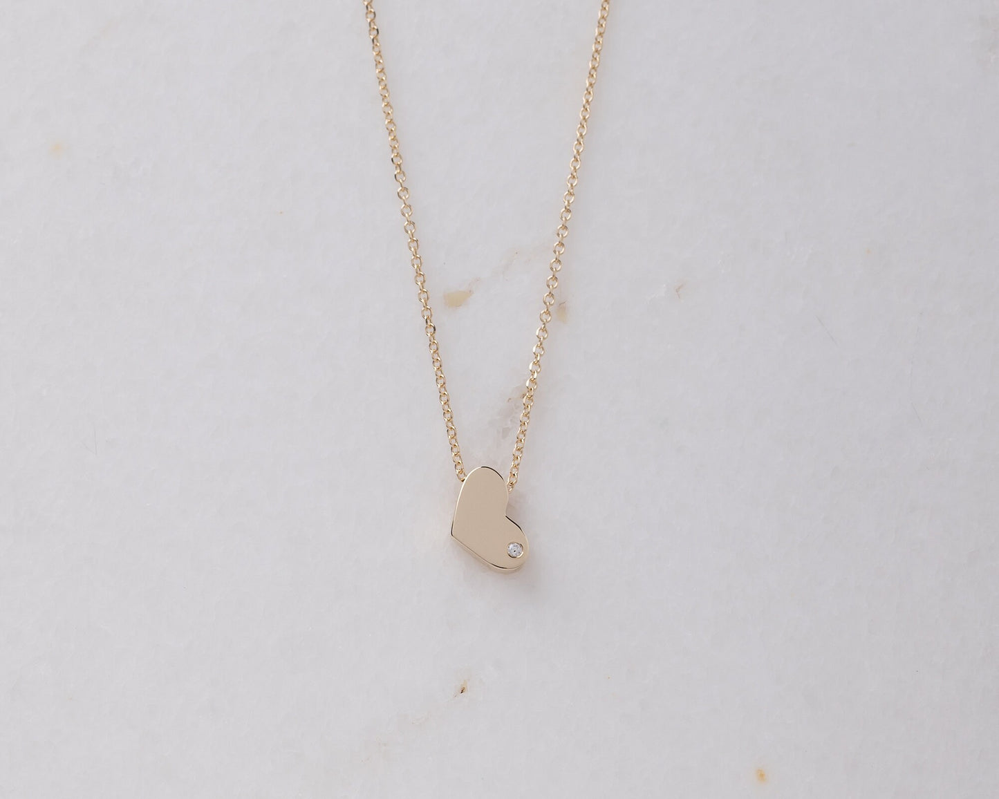 14k gold heart necklace with a modern, slightly asymmetrical design, with smooth, polished edges. A small diamond is embedded at the bottom right corner