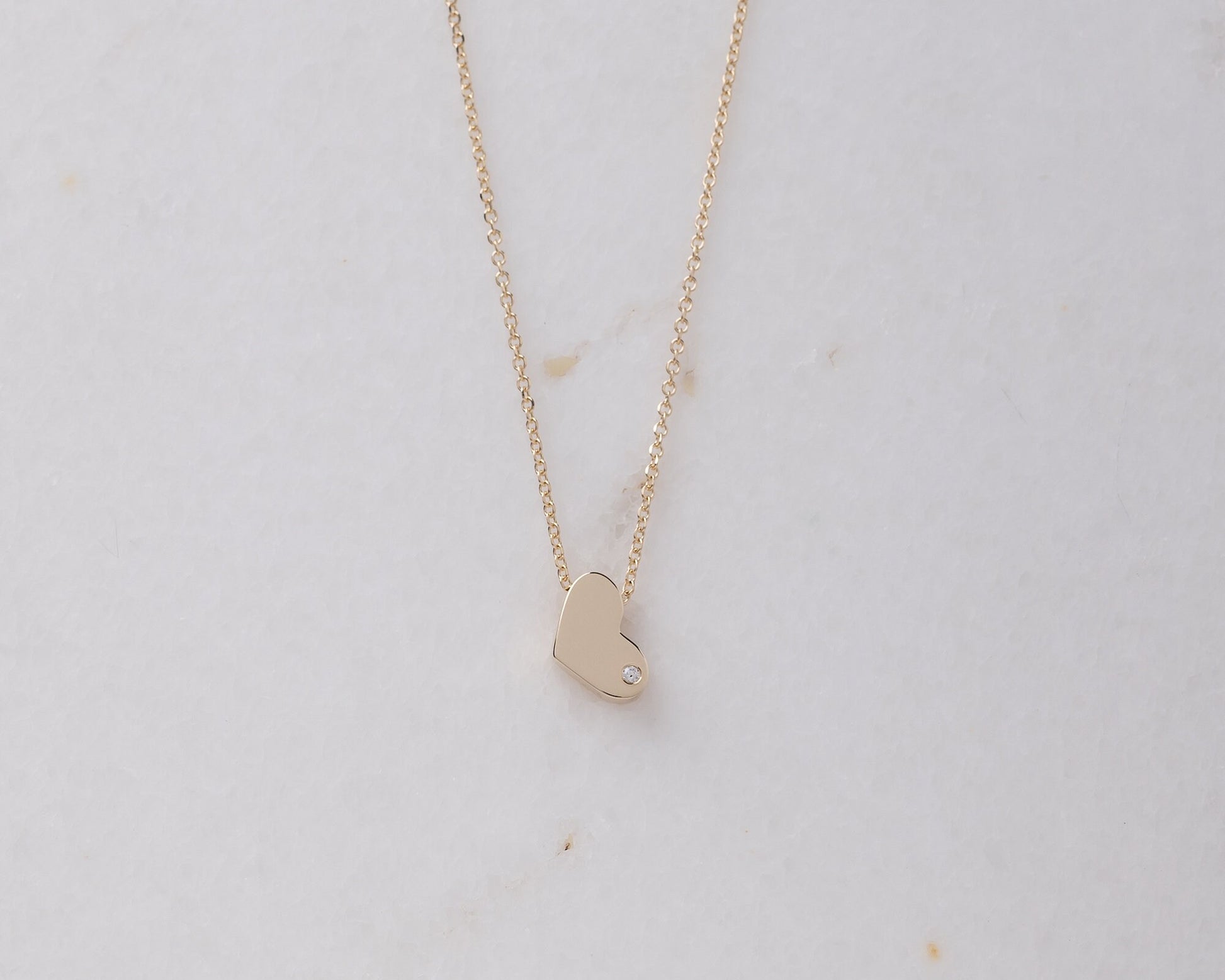 14k gold heart necklace with a modern, slightly asymmetrical design, with smooth, polished edges. A small diamond is embedded at the bottom right corner