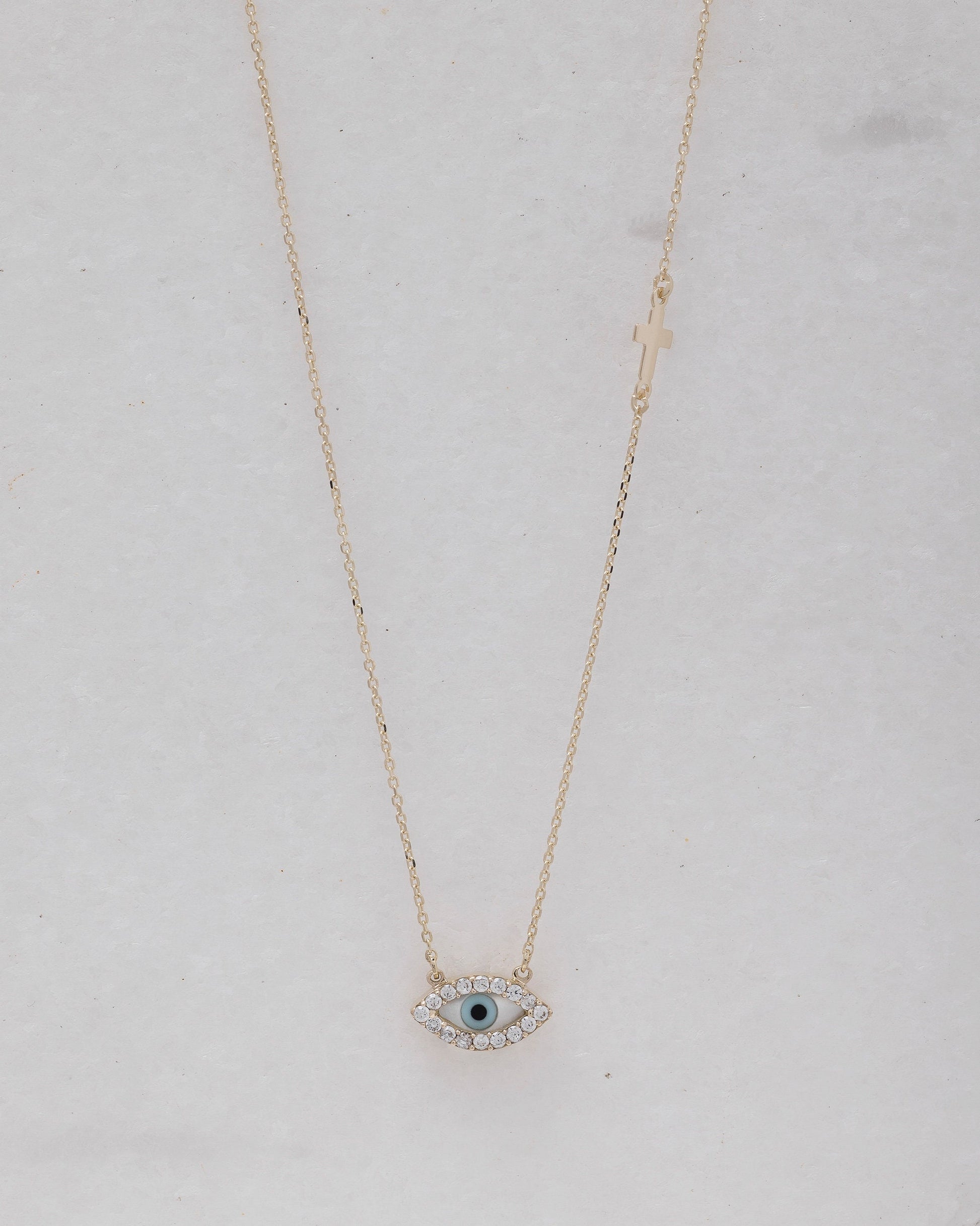 An oval-shaped mother of pearl evil eye necklace for women, crafted in 14k gold and adorned with cubic zirconia stones and a small side cross 