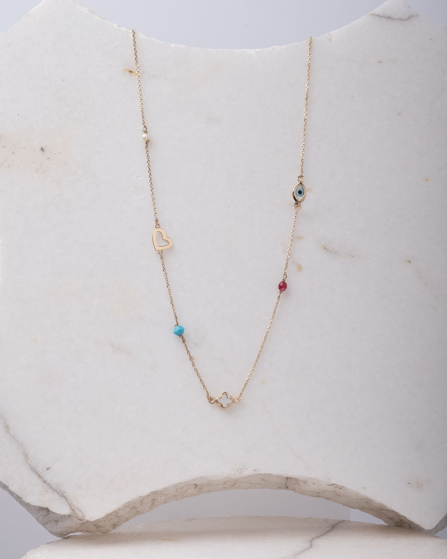 A station necklace featuring an evil eye, clover, heart, tourmaline, pearl, and turquoise stone set in 14k gold.