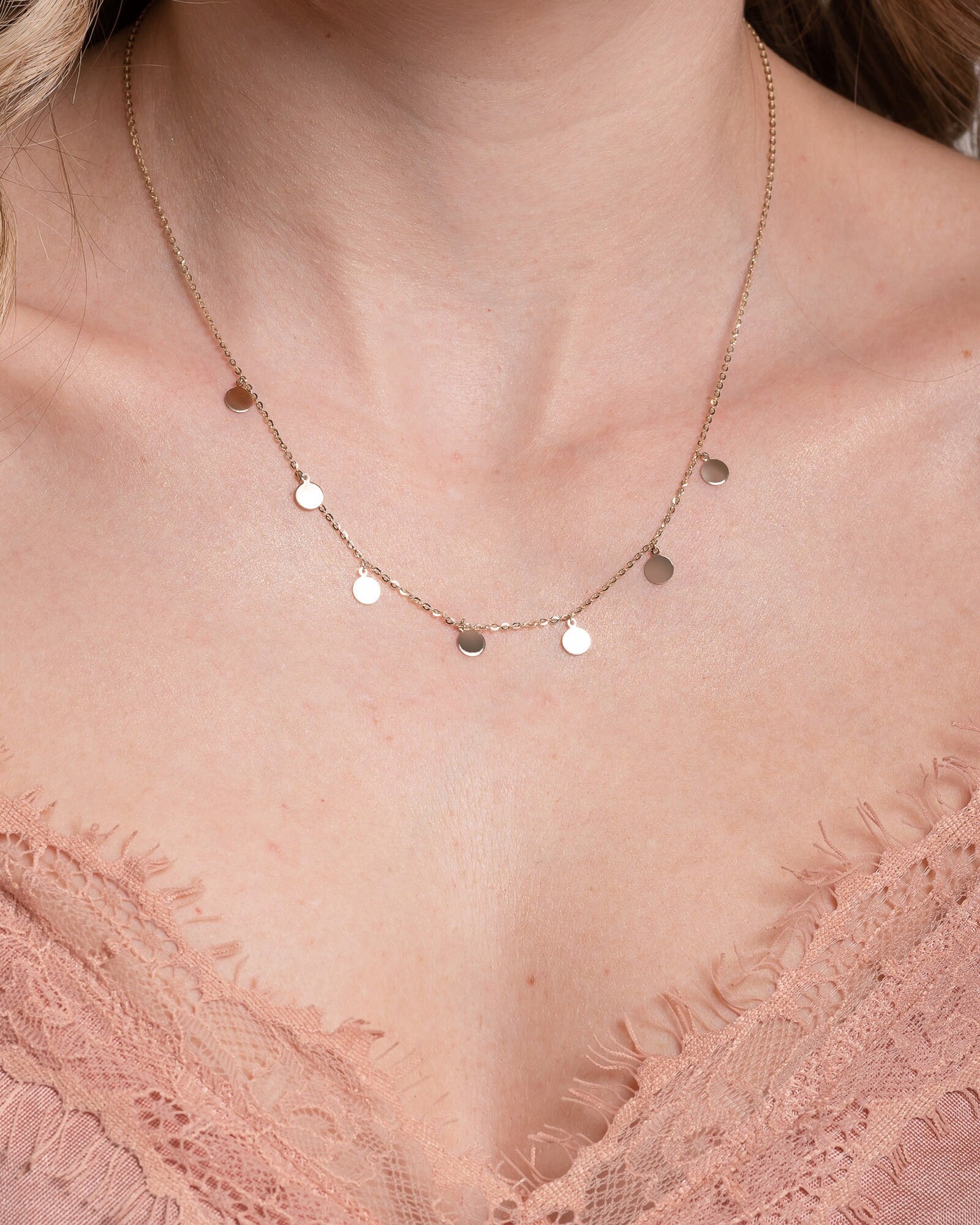 The photo shows a delicate gold necklace featuring multiple small, round, flat coin-shaped pendants evenly spaced along a fine gold chain. 