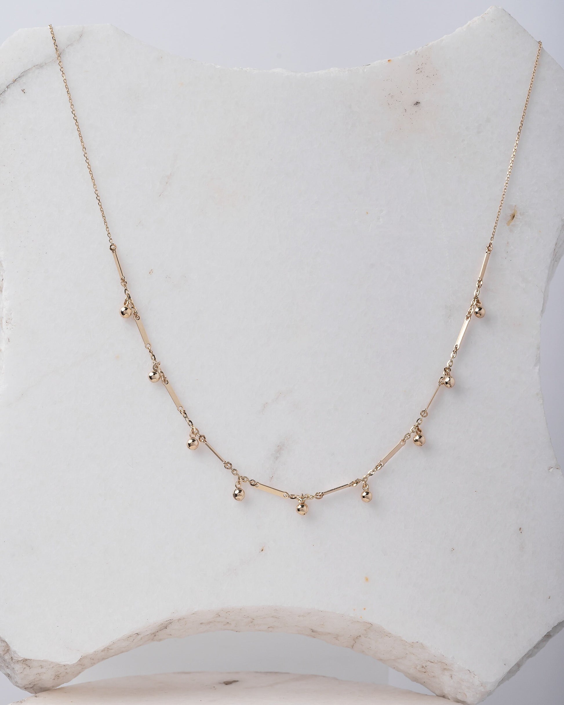 The photo shows a gold necklace featuring small, shiny, spherical bead-like pendants spaced along a fine chain. Between the beads, there are slim, rectangular bar accents.