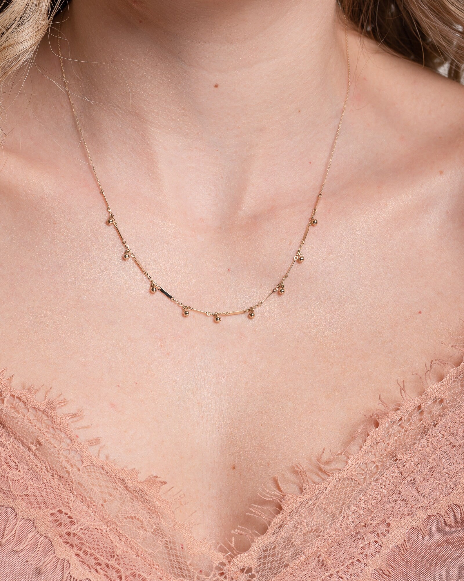 The photo shows a gold necklace featuring small, shiny, spherical bead-like pendants spaced along a fine chain. Between the beads, there are slim, rectangular bar accents.