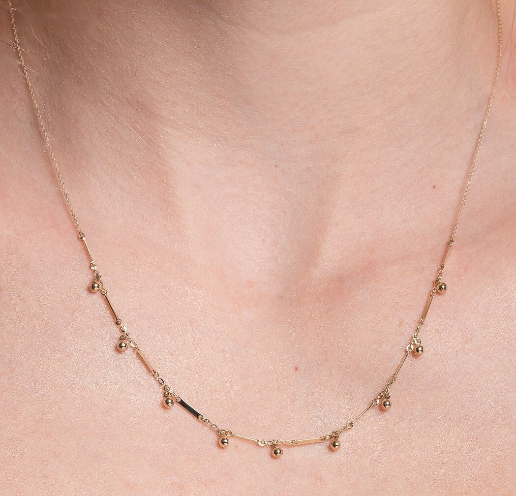 The photo shows a gold necklace featuring small, shiny, spherical bead-like pendants spaced along a fine chain. Between the beads, there are slim, rectangular bar accents.