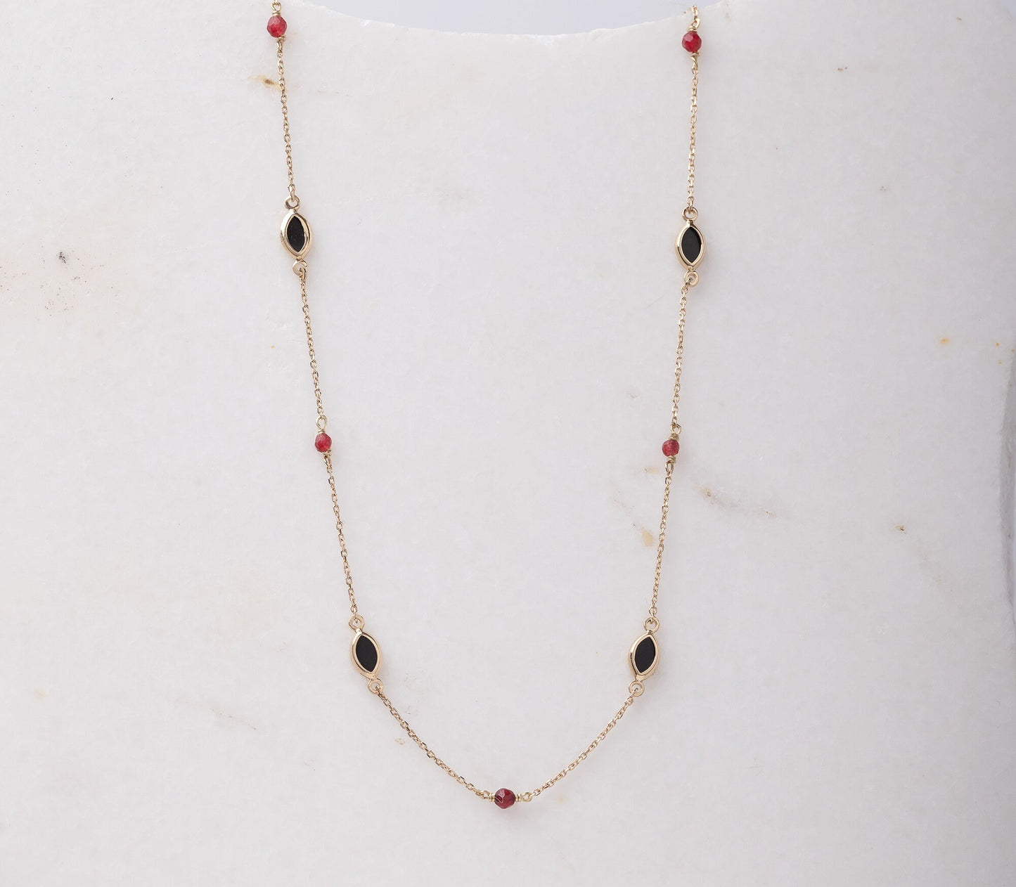 The photo shows a delicate gold necklace featuring small tourmaline gemstones and black marquise-shaped onyx accents, spaced along a fine chain. The contrasting colors create a striking and elegant design