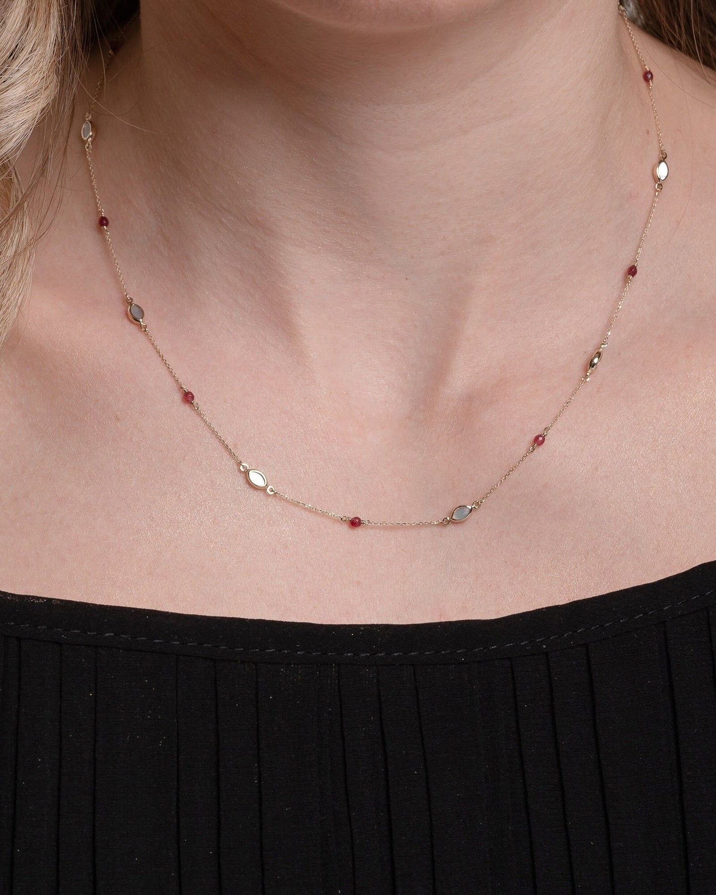 The photo shows a delicate gold necklace featuring small tourmaline gemstones and black marquise-shaped mother of pearl accents, spaced along a fine chain. The contrasting colors create a striking and elegant design