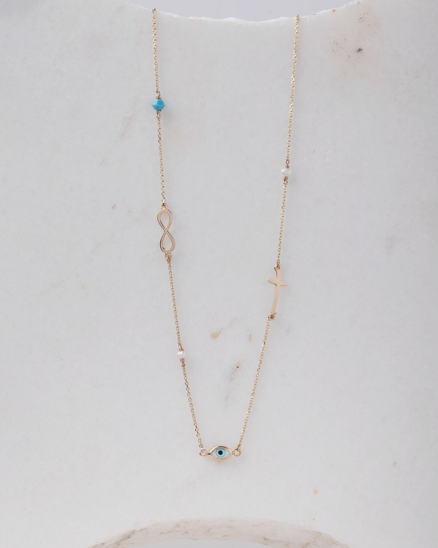 Station necklace featuring an evil eye, a cross, an infinity symbol, turquoise, and pearls in 14k gold.