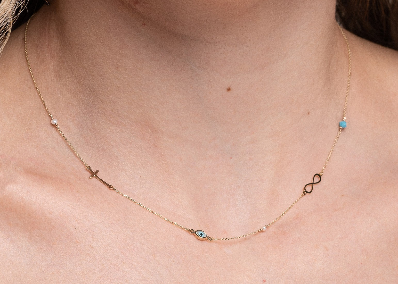 Station necklace featuring an evil eye, a cross, an infinity symbol, turquoise, and pearls in 14k gold.