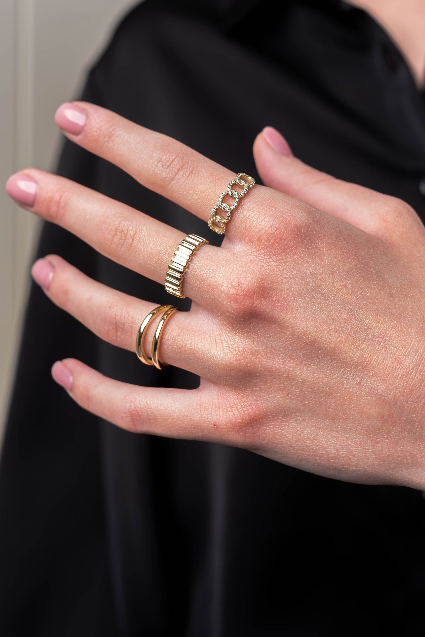 The image shows a hand wearing three elegant gold rings. One ring has a chain-link design with small diamonds, another features vertical bar details, and the third is a sleek double-band ring.