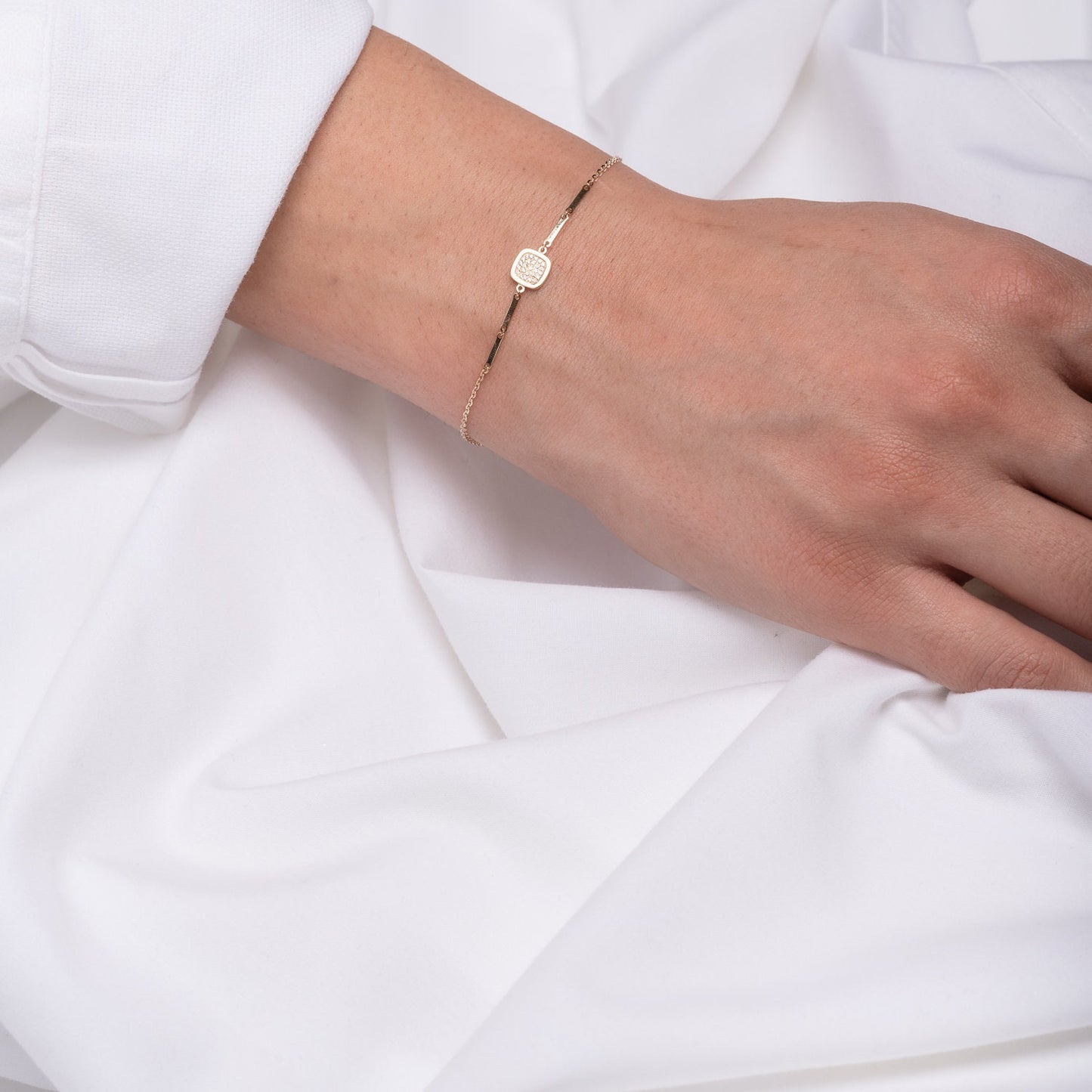 a hand wearing 14k gold charm bracelet with a square charm paved with white cubic zirconia stones