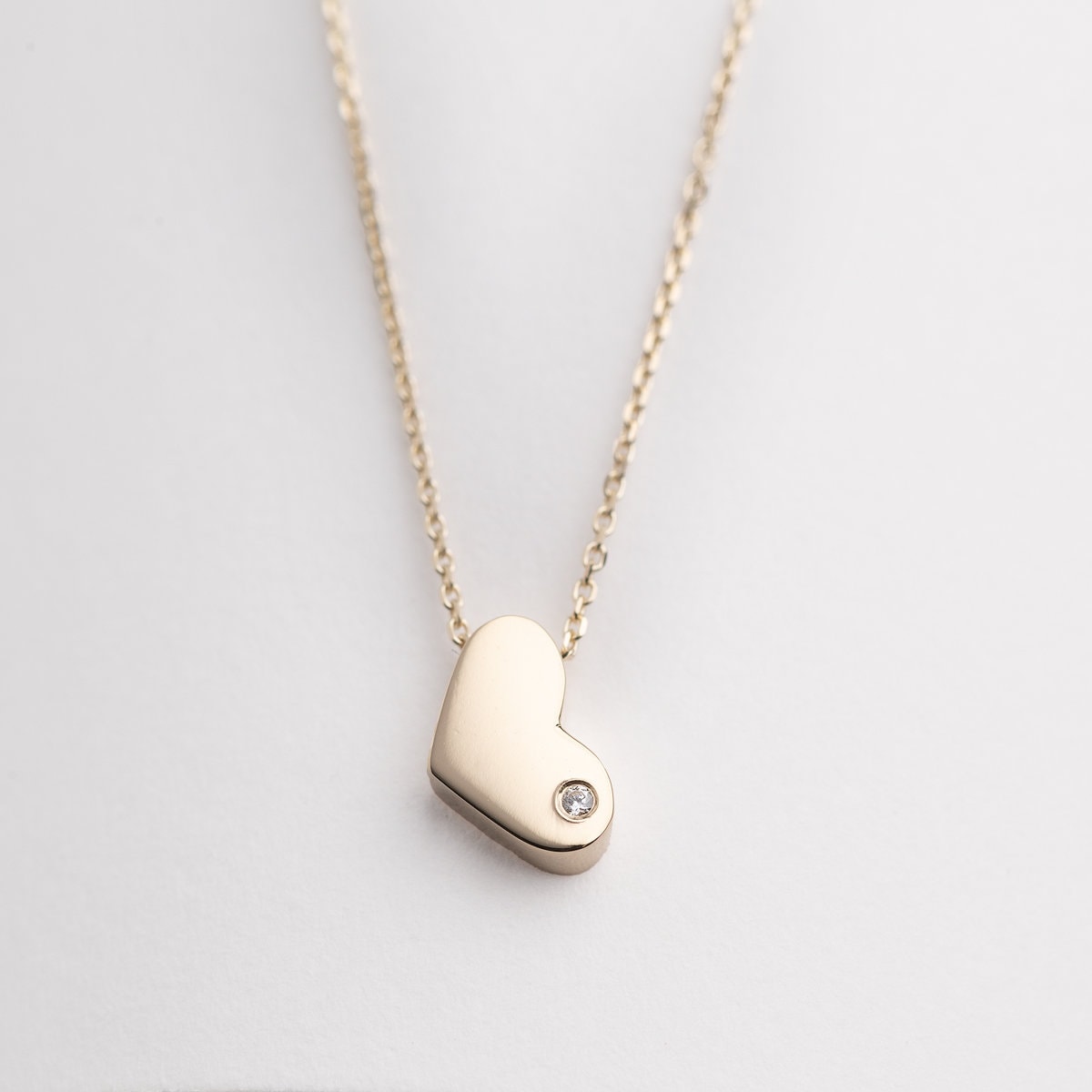 14k gold heart necklace with a modern, slightly asymmetrical design, with smooth, polished edges. A small diamond is embedded at the bottom right corner