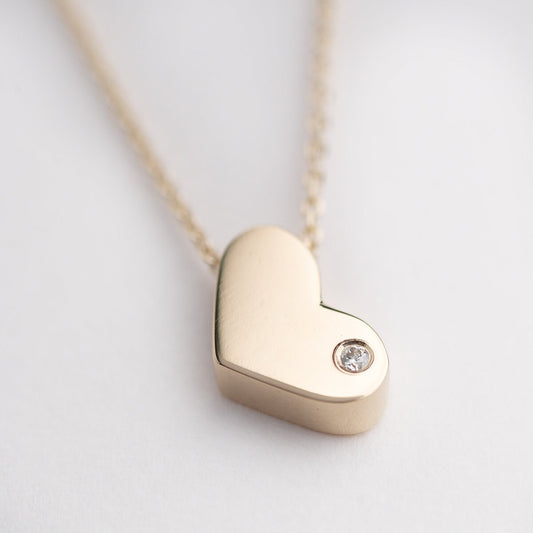 14k gold heart necklace with a modern, slightly asymmetrical design, with smooth, polished edges. A small diamond is embedded at the bottom right corner