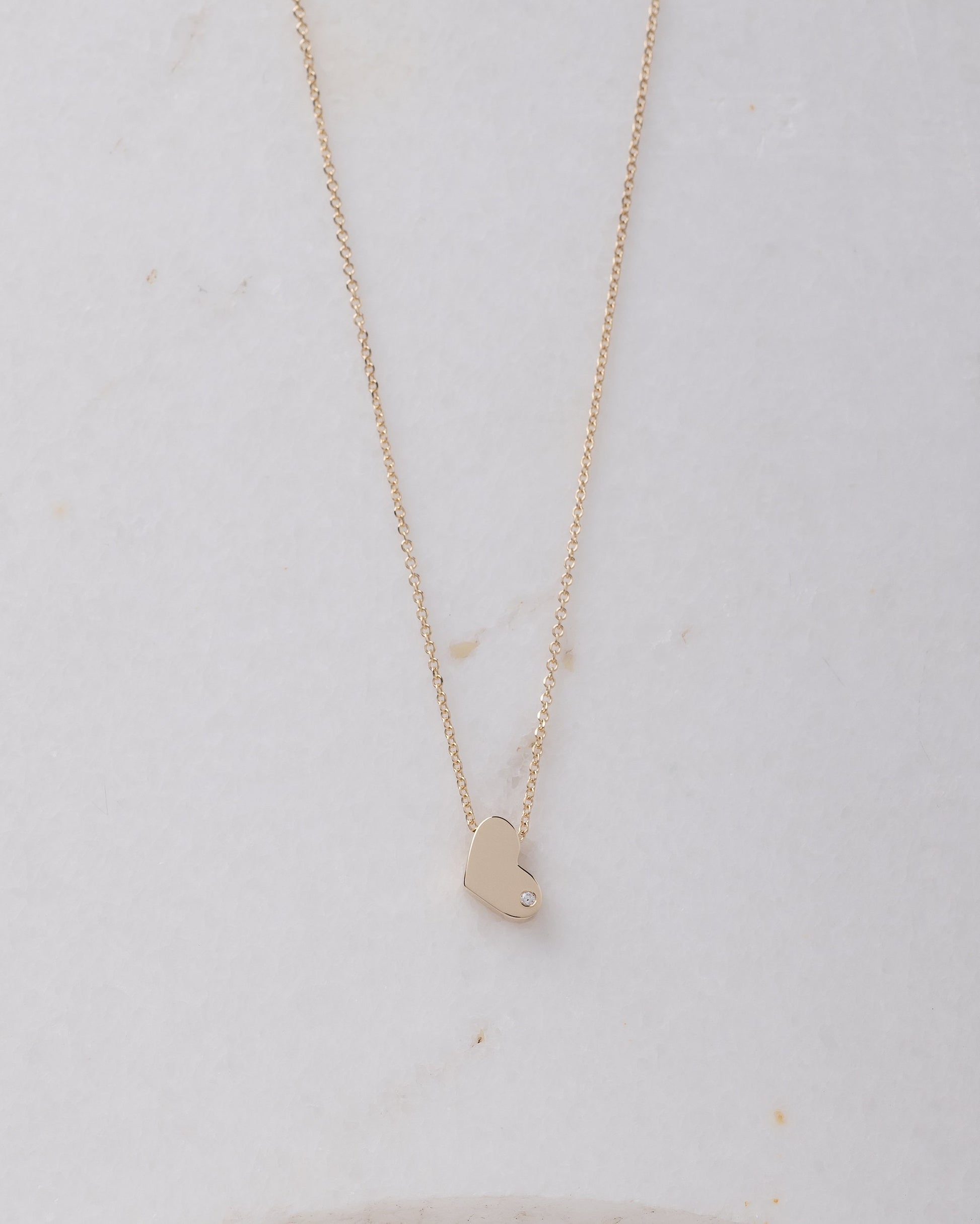 14k gold heart necklace with a modern, slightly asymmetrical design, with smooth, polished edges. A small diamond is embedded at the bottom right corner