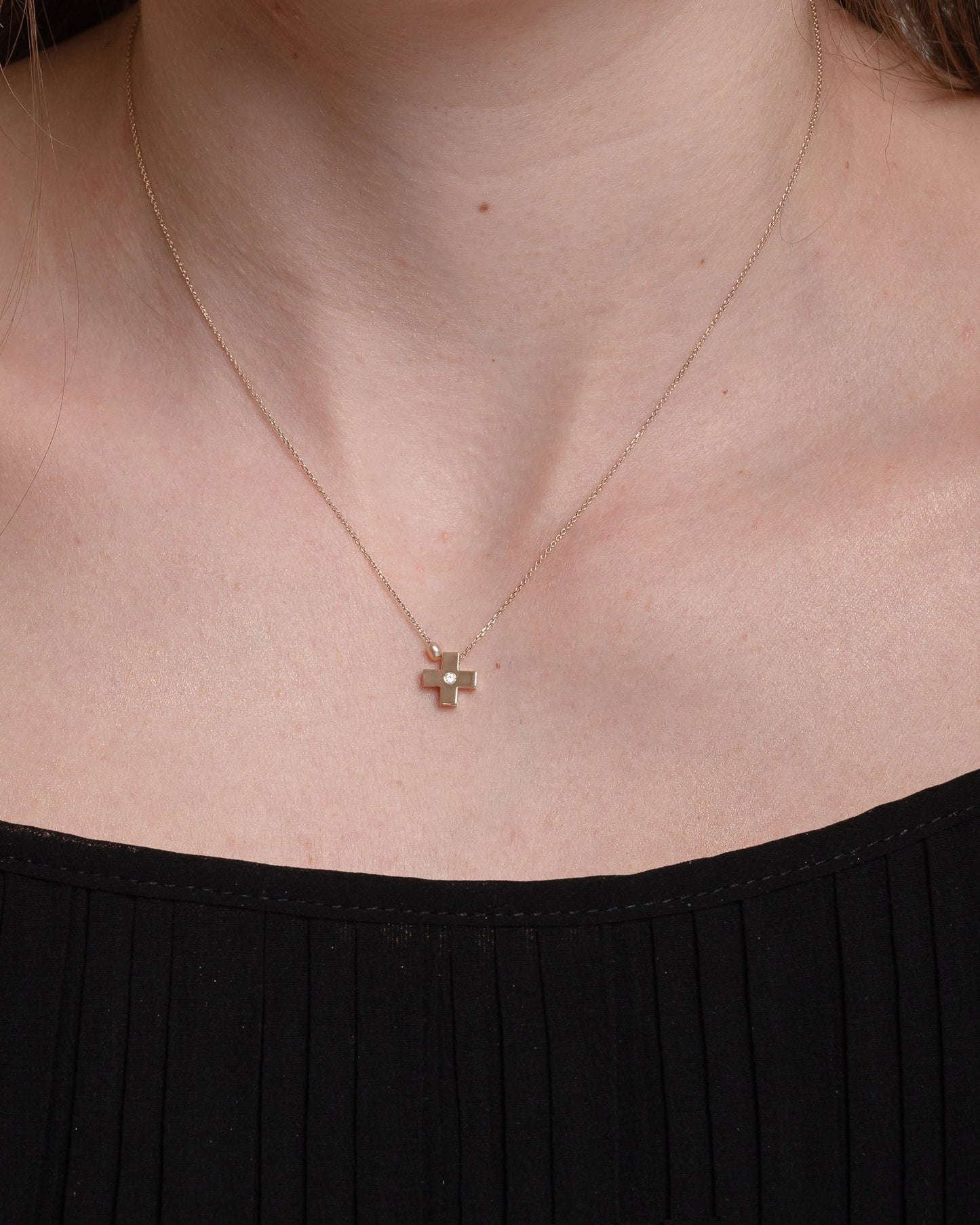 small diamond cross necklace with a small pearl in 14k yellow gold for women worn by a model 