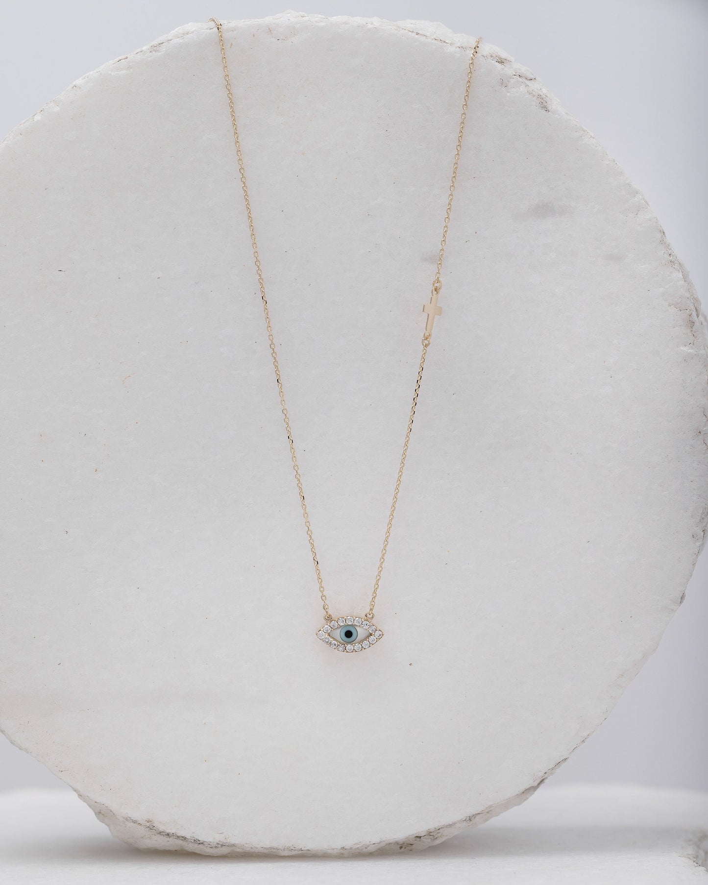 An oval-shaped mother of pearl evil eye necklace for women, crafted in 14k gold and adorned with cubic zirconia stones and a small side cross 