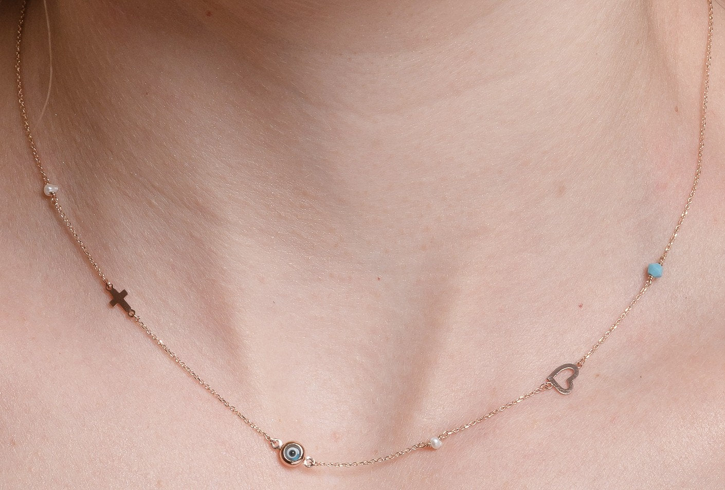 A station necklace featuring an evil eye, heart, cross, pearls, and a turquoise stone, all crafted in 14k gold.