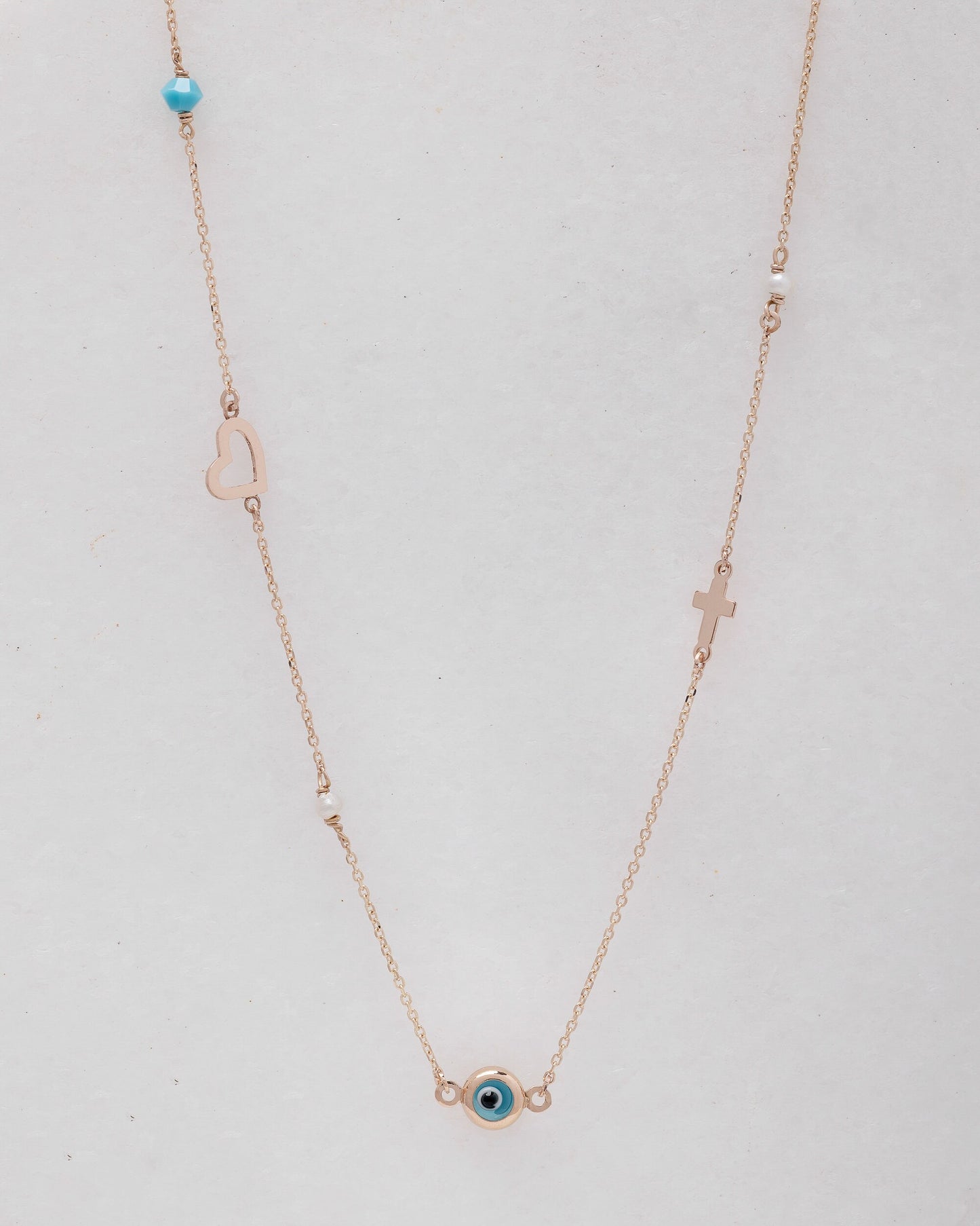 A station necklace featuring an evil eye, heart, cross, pearls, and a turquoise stone, all crafted in 14k gold.