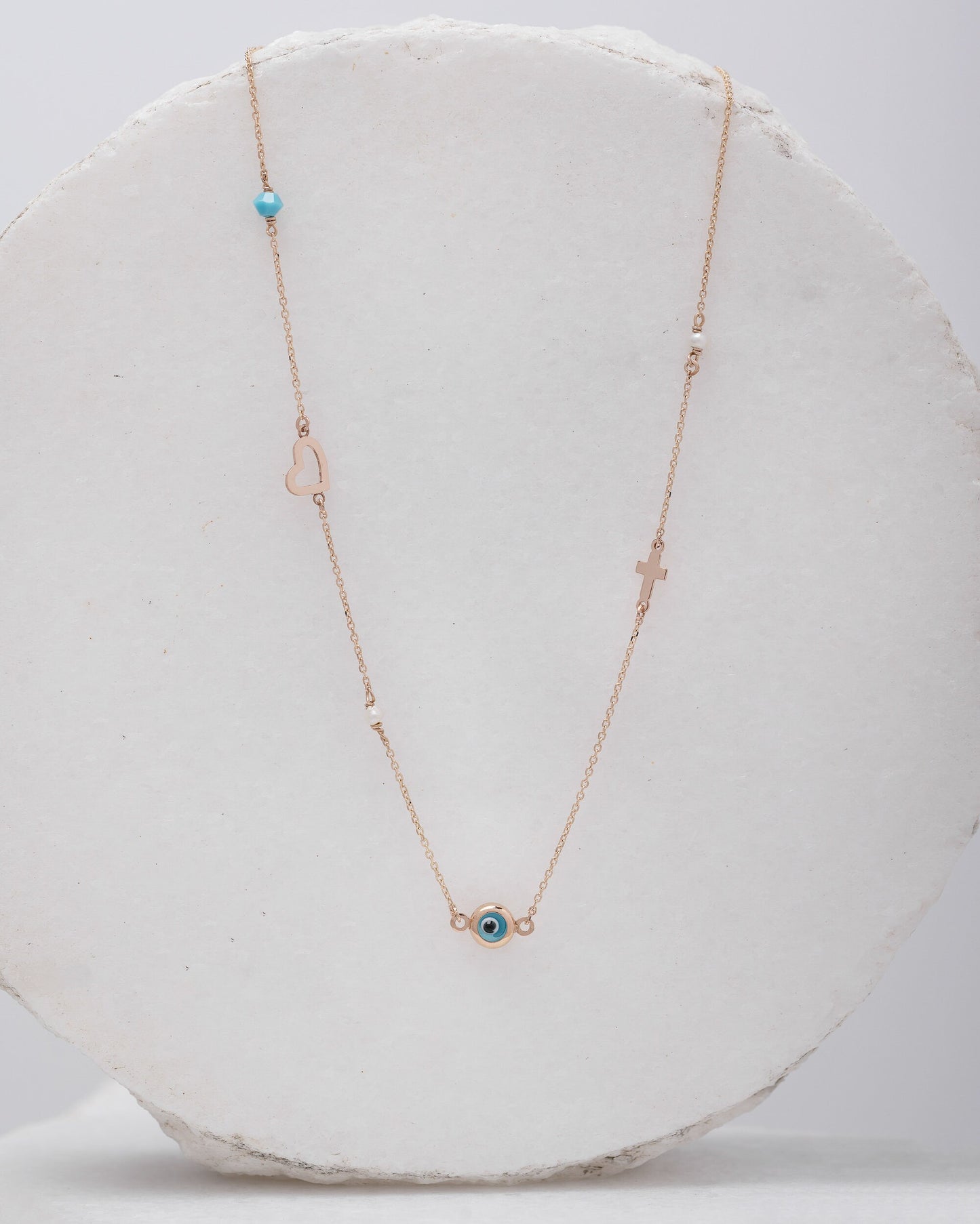 A station necklace featuring an evil eye, heart, cross, pearls, and a turquoise stone, all crafted in 14k gold.