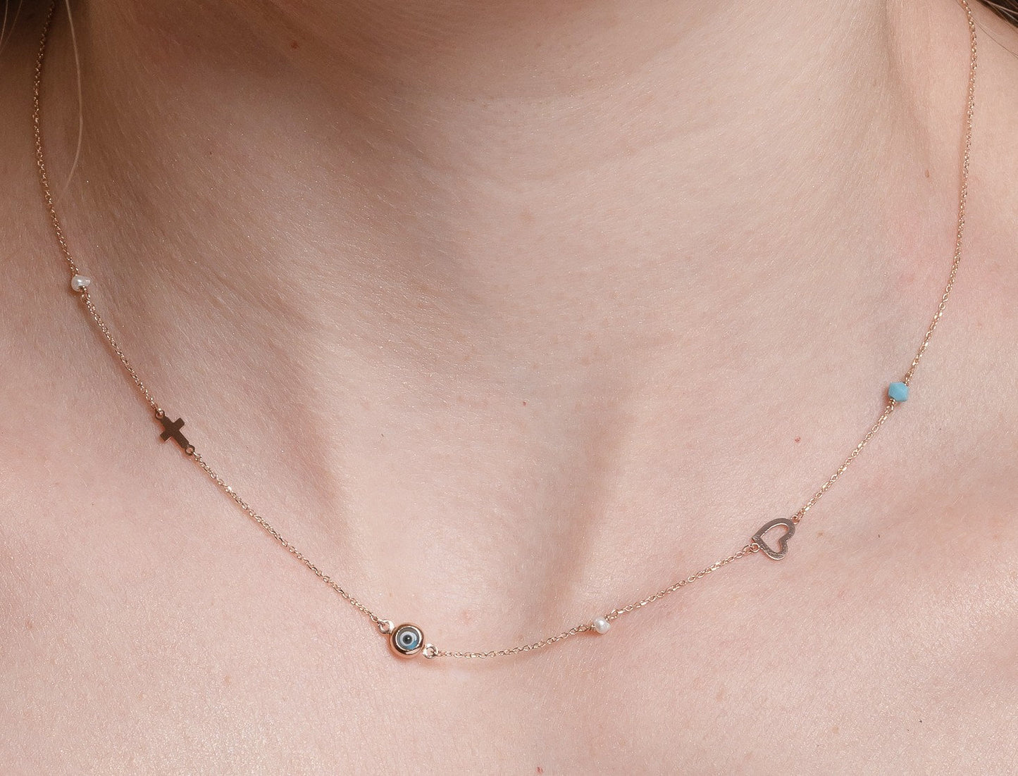 A station necklace featuring an evil eye, heart, cross, pearls, and a turquoise stone, all crafted in 14k gold.