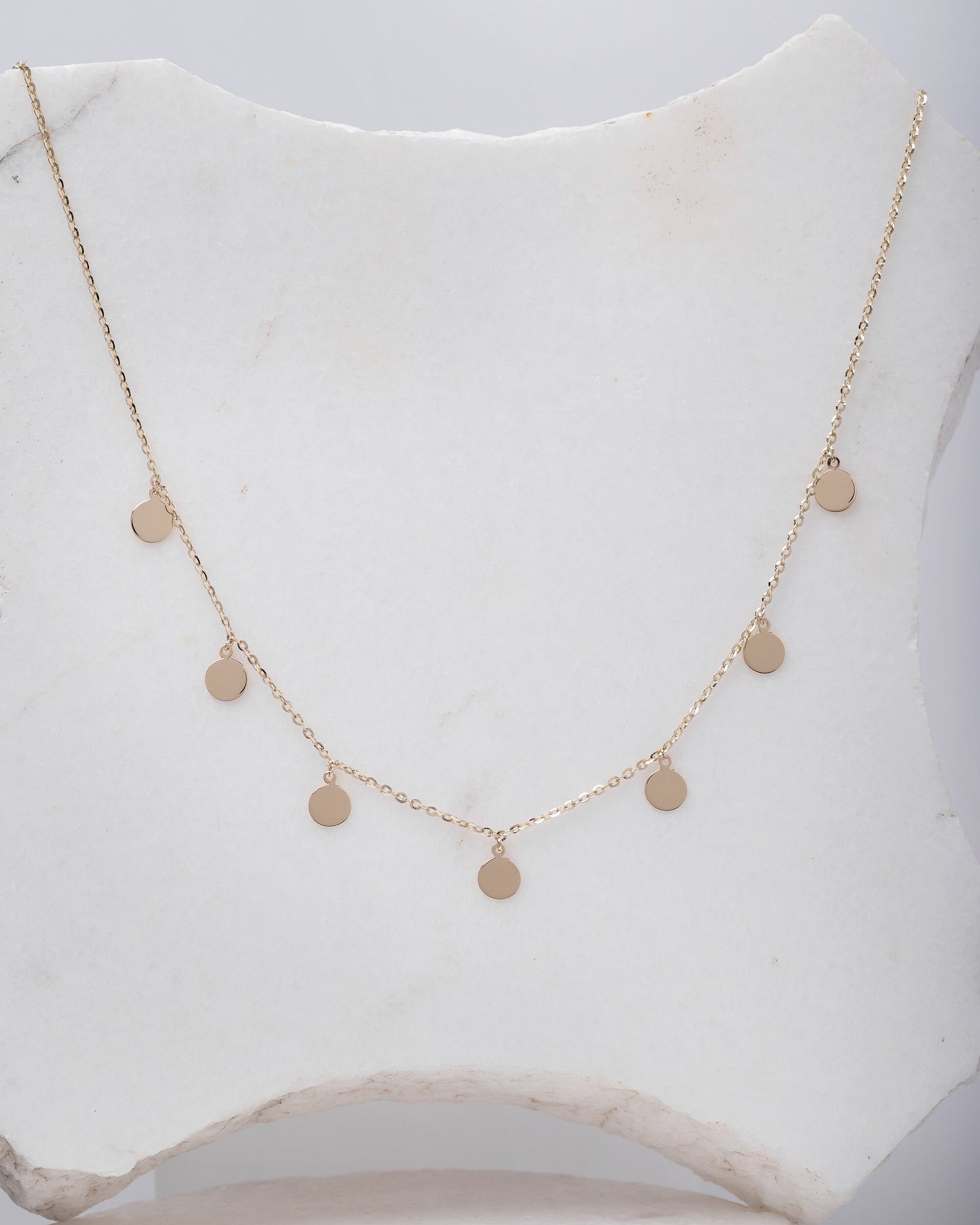 The photo shows a delicate gold necklace featuring multiple small, round, flat coin-shaped pendants evenly spaced along a fine gold chain. 