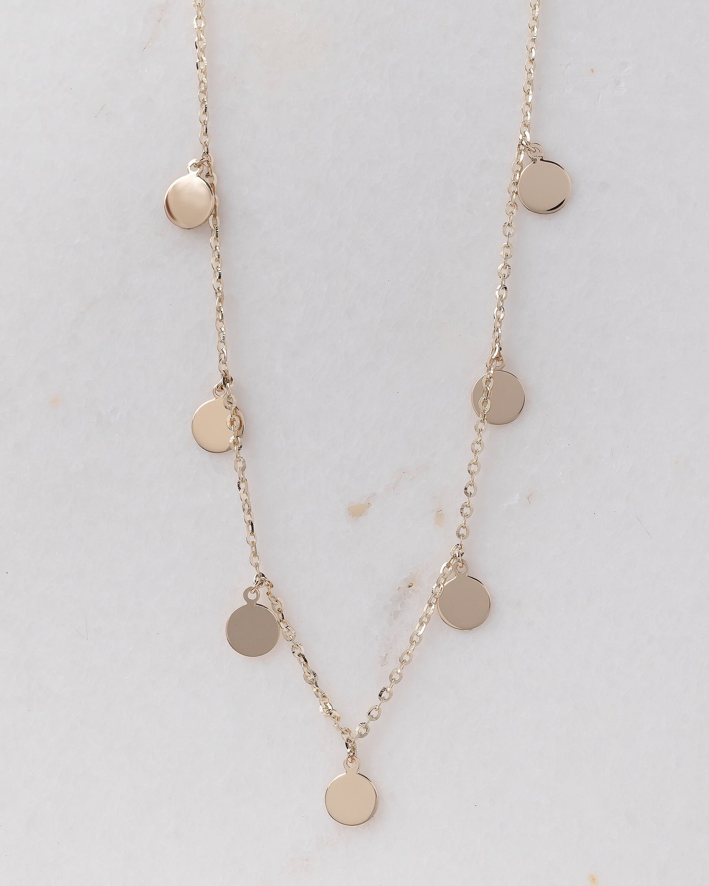 The photo shows a delicate gold necklace featuring multiple small, round, flat coin-shaped pendants evenly spaced along a fine gold chain. 